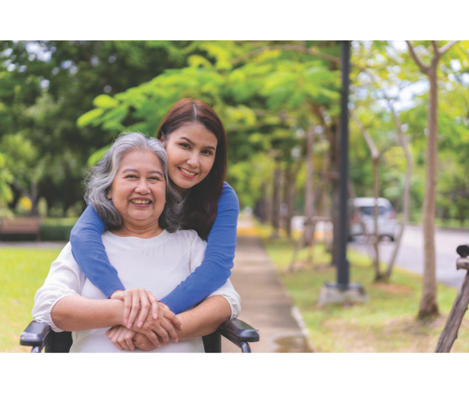 Young woman with dark brown hair has her arms around an elderly woman in a wheelchair, outside with trees in back ground